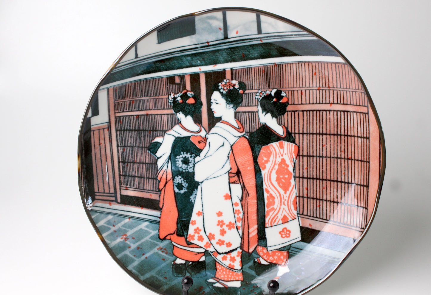 Porcelain dining plate with a soft wavy edge, featuring an etched illustration of three geishas in vibrant kimonos standing before a traditional Japanese backdrop.
