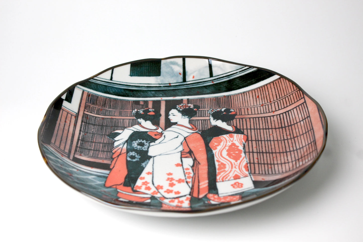 Round ceramic plate with a softly wavy edge, featuring an etched illustration of three geishas in vibrant kimonos standing before a traditional Japanese backdrop.