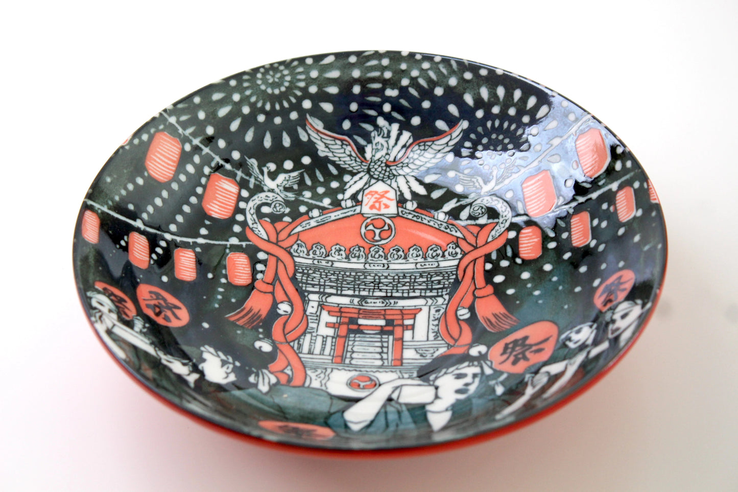 A handcrafted Japanese ceramic plate featuring a vibrant festival scene with red lanterns, a traditional shrine, and a phoenix illustration against a dark background with firework patterns on the inside. The intricate design captures the essence of a lively Matsuri (festival), blending cultural heritage with functional artistry.