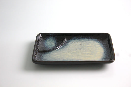 A handcrafted ceramic snack plate featuring a glossy midnight black and moonlit ivory glaze, with an integrated sauce section, perfect for serving sushi and appetizers.