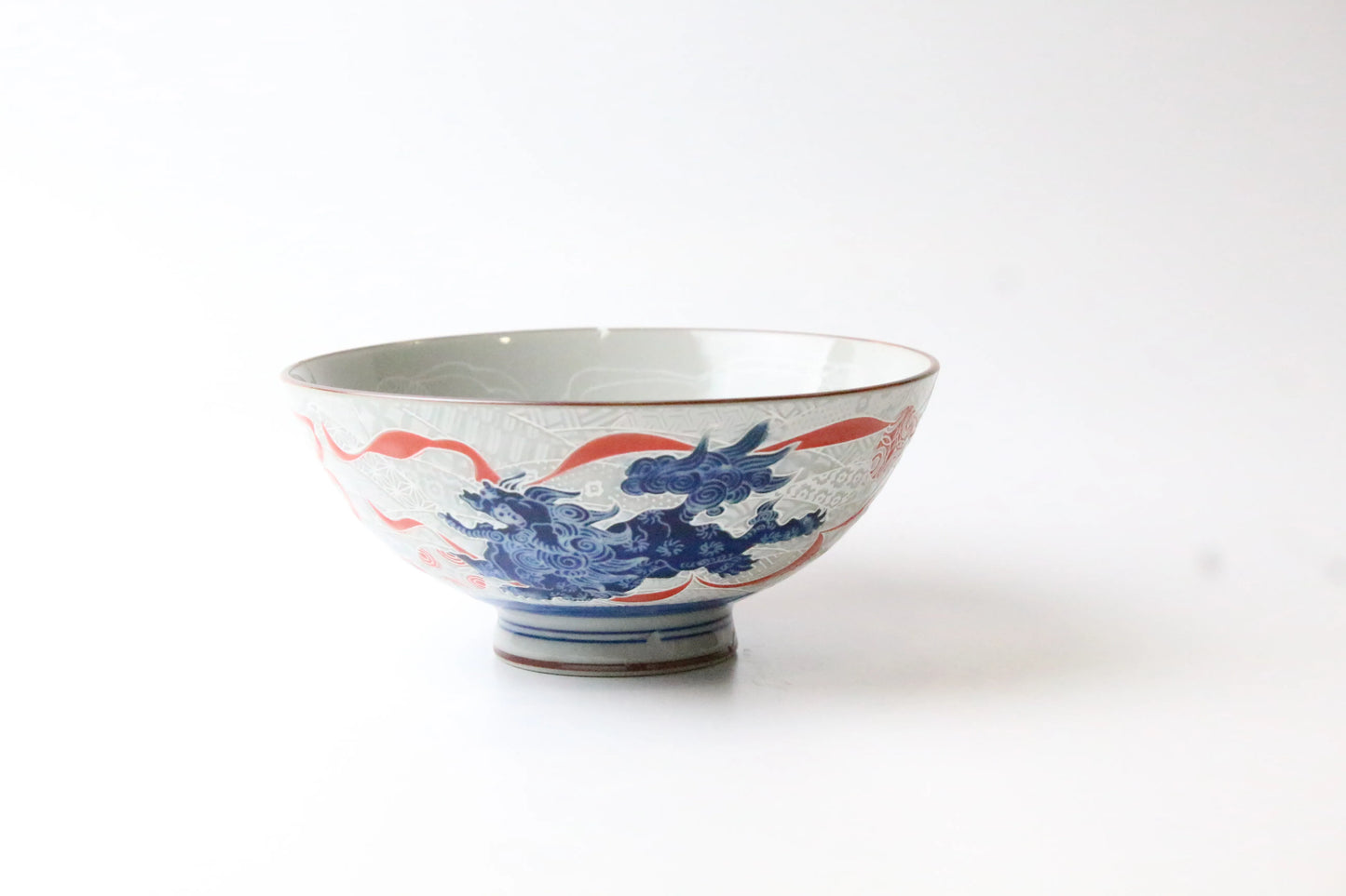 A handcrafted porcelain rice bowl featuring an intricately etched Japanese lion (Shishi) in blue and red on the exterior, with a glossy finish and a printed lion design inside.