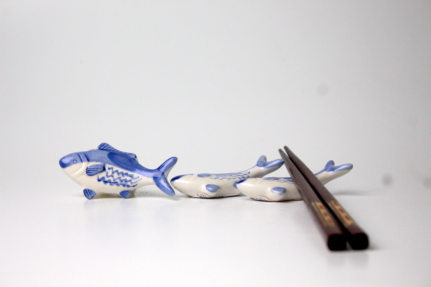 A set of ceramic tuna-shaped spoon rests in blue and white, accompanied by wooden chopsticks, displayed on a white surface.