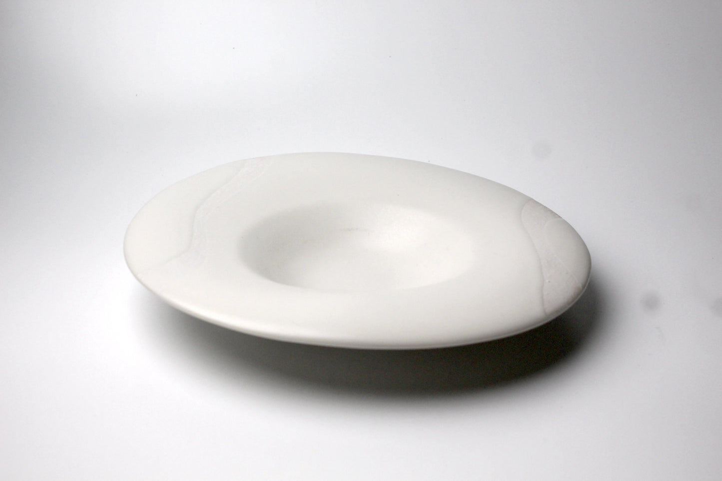 A shallow ceramic plate with a white matte finish, featuring a subtle textured surface and soft, natural ivory tones.