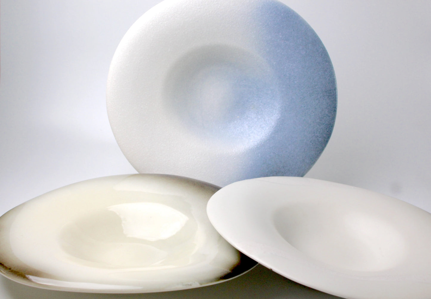 A set of handcrafted shallow ceramic plates with a mix of glossy and matte finishes, featuring cool glazing and a textural surface in soft, natural hues.