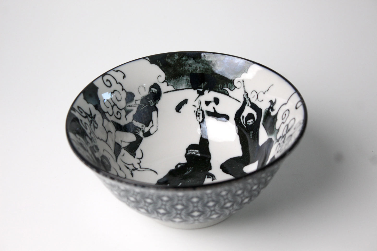 A black-and-white ceramic ramen bowl featuring dynamic ninja figures and swirling clouds, set against a white interior.