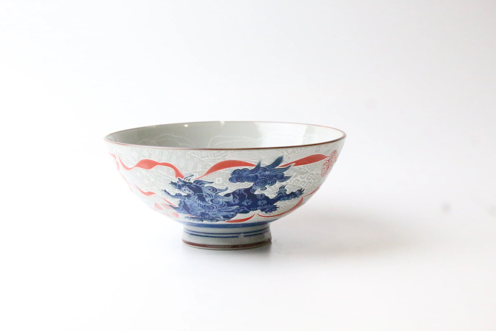 A handcrafted porcelain rice bowl featuring an intricately etched Japanese lion (Shishi) in blue and red on the exterior, with a glossy finish and a printed lion design inside.