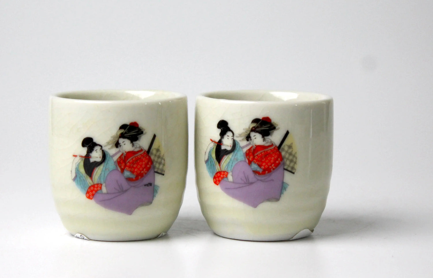 Two ceramic sake cups with decorative designs of Japanese women on an off-white background
