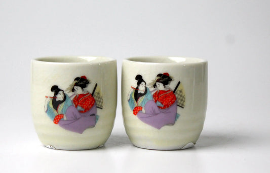 Two ceramic sake cups with decorative designs of Japanese women on an off-white background