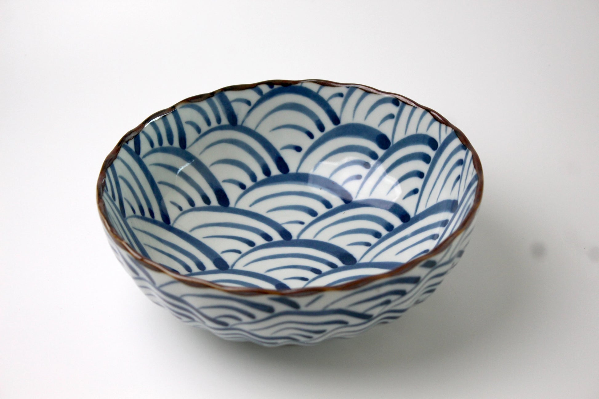 A Japanese ceramic bowl featuring waves of the blue sea pattern in deep blue, with a scalloped rim and a glossy finish.