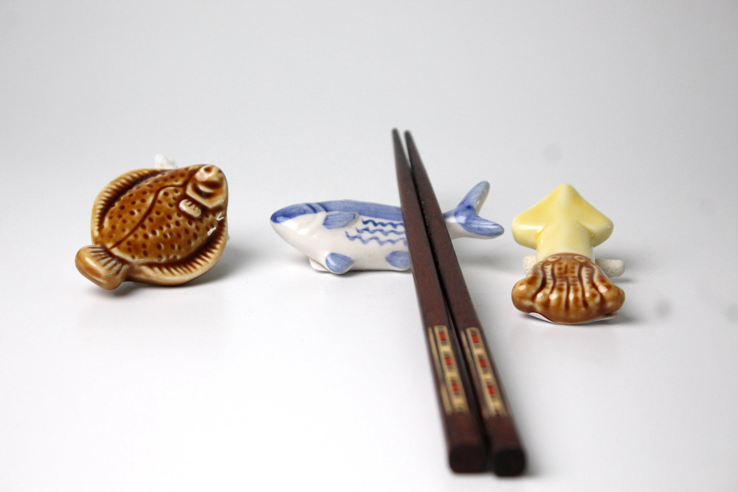 A set of three ceramic spoon rests shaped like a flounder, a tuna, and a squid, with a pair of wooden chopsticks resting on the squid spoon rest. The spoon rests are hand-painted with intricate details, set against a clean white background.